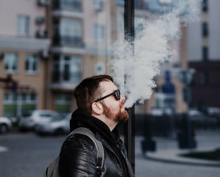 Man wearing sunglasses and a black leather jacket exhaling smoke outdoors in an urban UK setting.