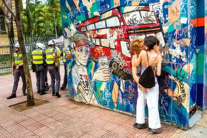 Candid street photo of two women embracing by colorful graffiti wall with police officers nearby in urban setting.
