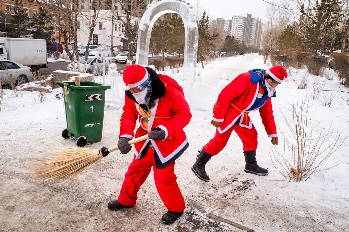 Two people dressed in red Santa outfits sweeping a snowy street in an urban winter setting, street photographer capture.