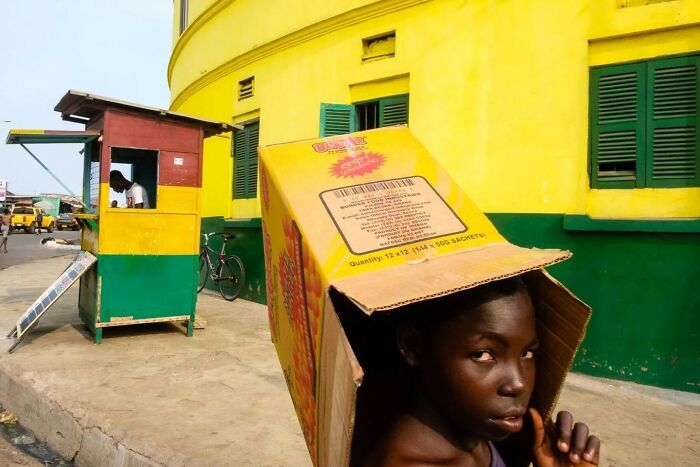 Boy with a cardboard box on his head captured by street photographer in vibrant urban setting with yellow and green buildings.
