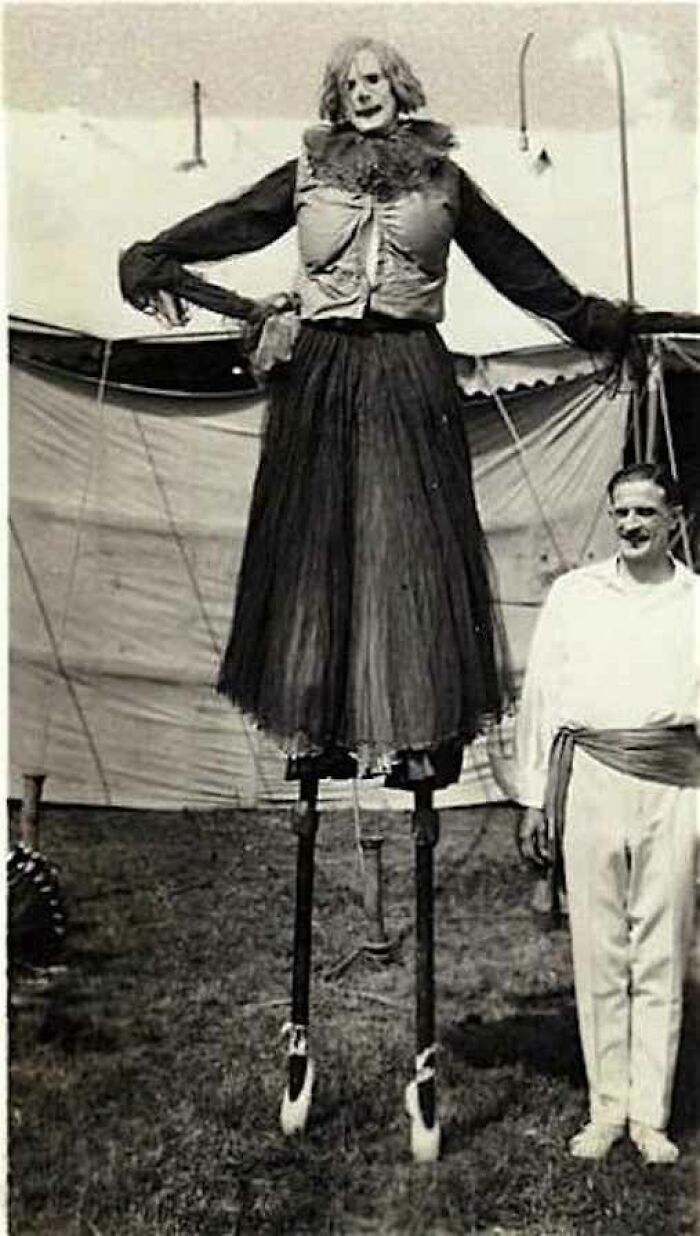 Black and white creepy vintage photo of a person on stilts dressed as a tall unsettling clown figure outdoors.