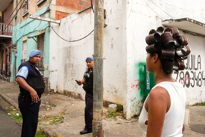 Two police officers stand on a street corner while a woman with hair rollers looks on in a candid street photographer shot.