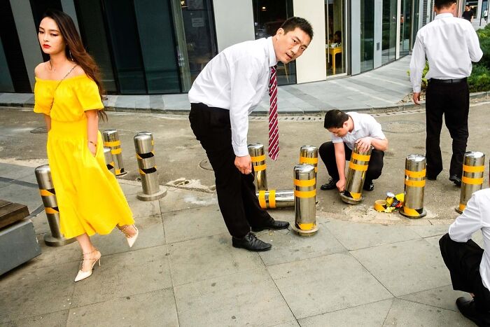 Street photographer captures candid moment of woman in yellow dress and men working with bollards on urban sidewalk.