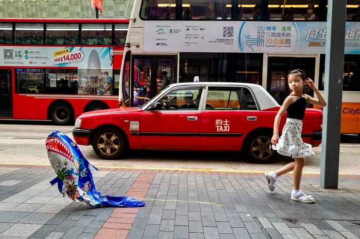 Young girl in a city street scene with red taxi and buses, captured by a street photographer’s candid moment.