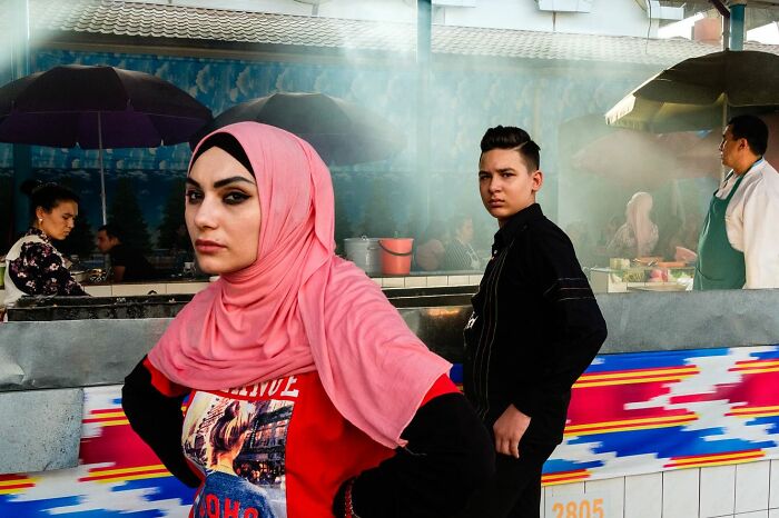 Street photographer captures candid moment of a woman in a pink hijab and a young man in an urban outdoor market.