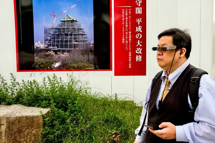 Man wearing futuristic glasses and holding a device, standing near a construction site in a candid street photography moment