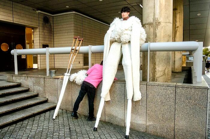 Street photographer captures person in unusual stilts costume and another leaning over a ledge in urban setting.