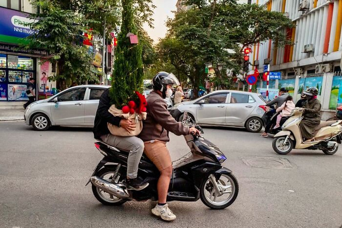 Two people on a scooter, one carrying a Christmas tree and red ornaments, captured by street photographer.