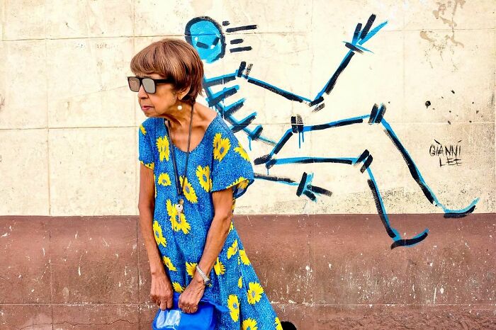 Woman in a blue sunflower dress posing candid street photo with playful wall art, captured by award-winning photographer.