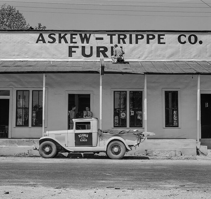 Colorized historical photo of a worker painting a sign on a bright yellow building with a vintage blue truck parked outside.