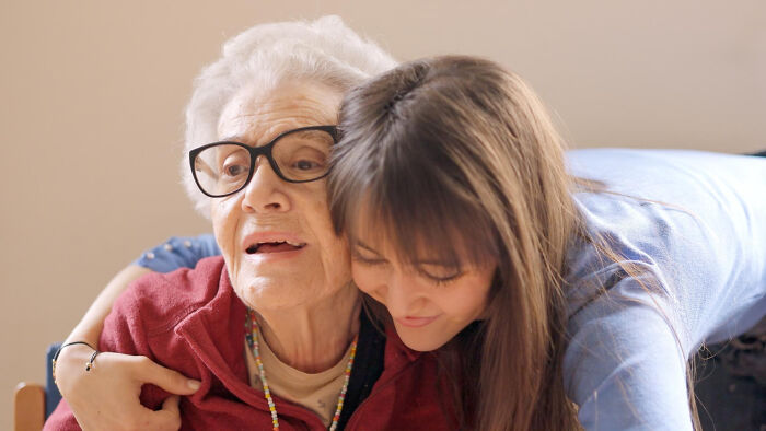 Elderly woman with glasses being warmly hugged by a younger woman, symbolizing a priceless reward of love and connection.