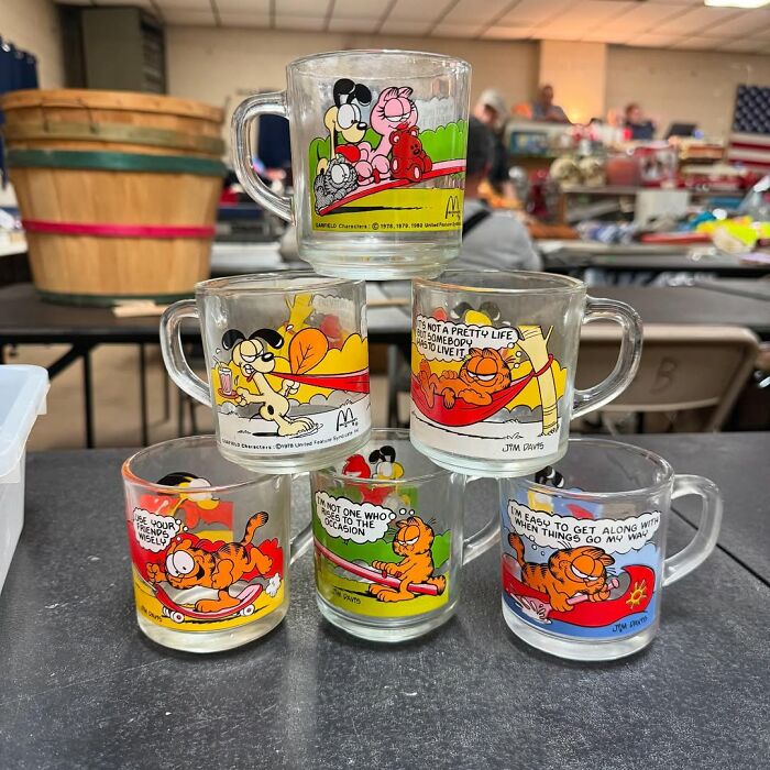 Stack of vintage Garfield-themed secondhand glass mugs displayed at a flea market with other items in the background.