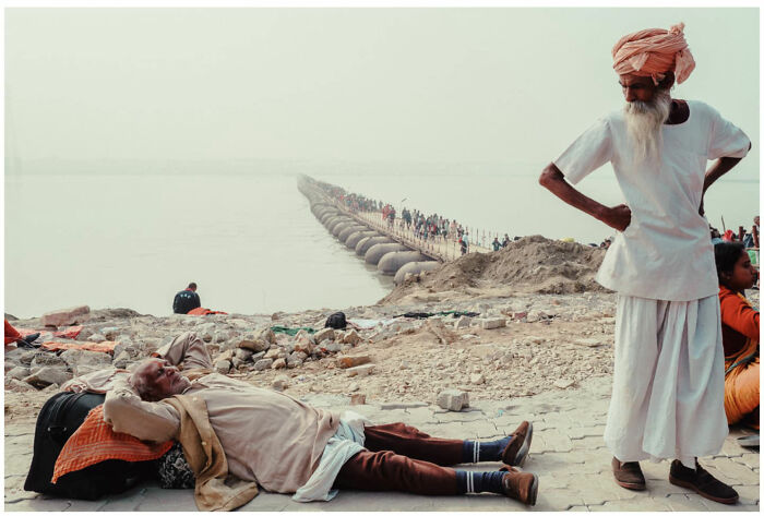 A man resting by the riverbank with a crowd walking over a floating bridge, capturing raw street moments in Asia.