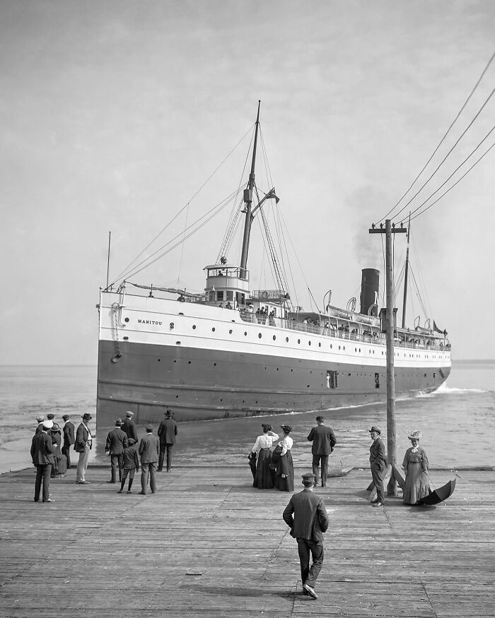 Colorized historical photo of a large red and white ship docked with people dressed in early 20th century clothing on the pier.