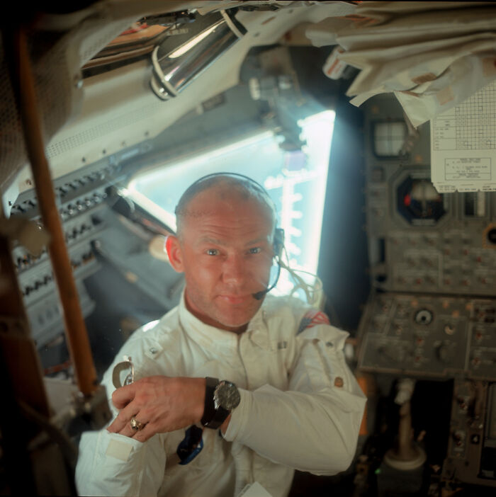 Astronaut inside spacecraft cockpit wearing headset and white suit, surrounded by control panels and instruments.