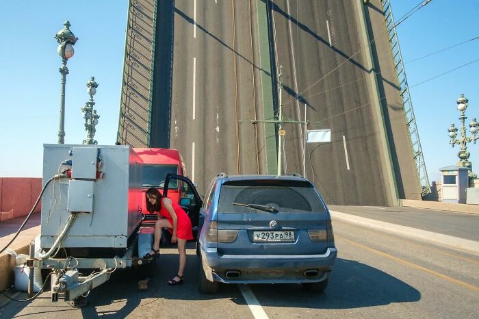 Street photographer captures woman in red dress exiting car on raised drawbridge with ornate street lamps in background.