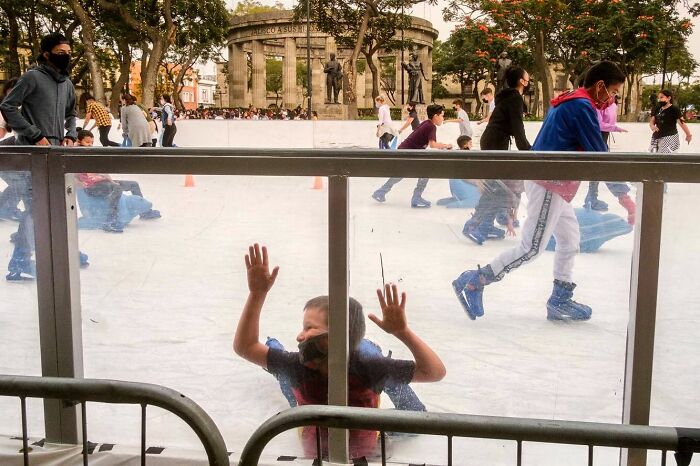 Child pressing hands on glass at outdoor ice rink with street photographer capturing candid moments of daily life
