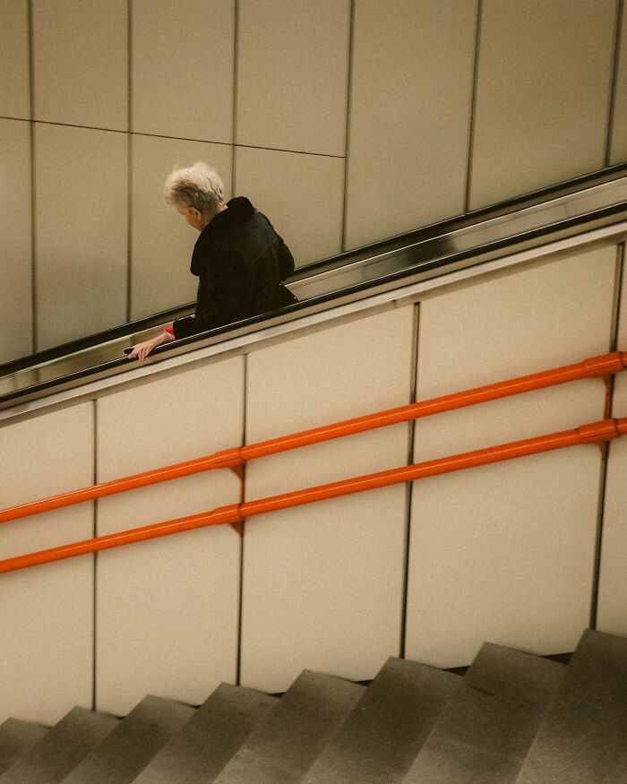 Elderly person in black coat on escalator with orange handrails in urban street photography style by Helsinki photographer