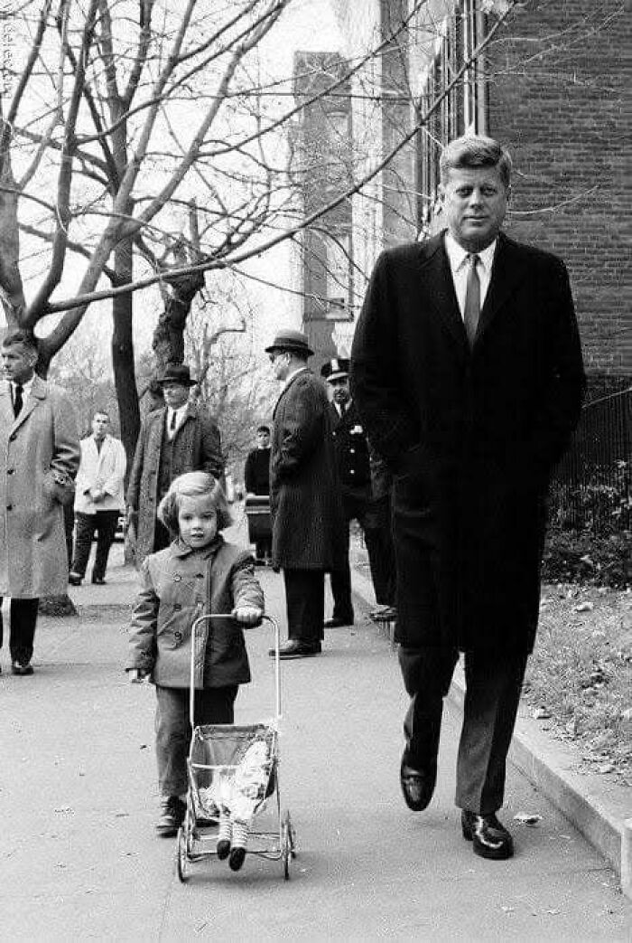 John F. Kennedy walking with a child pushing a doll stroller in a fascinating historical photo on a city sidewalk.