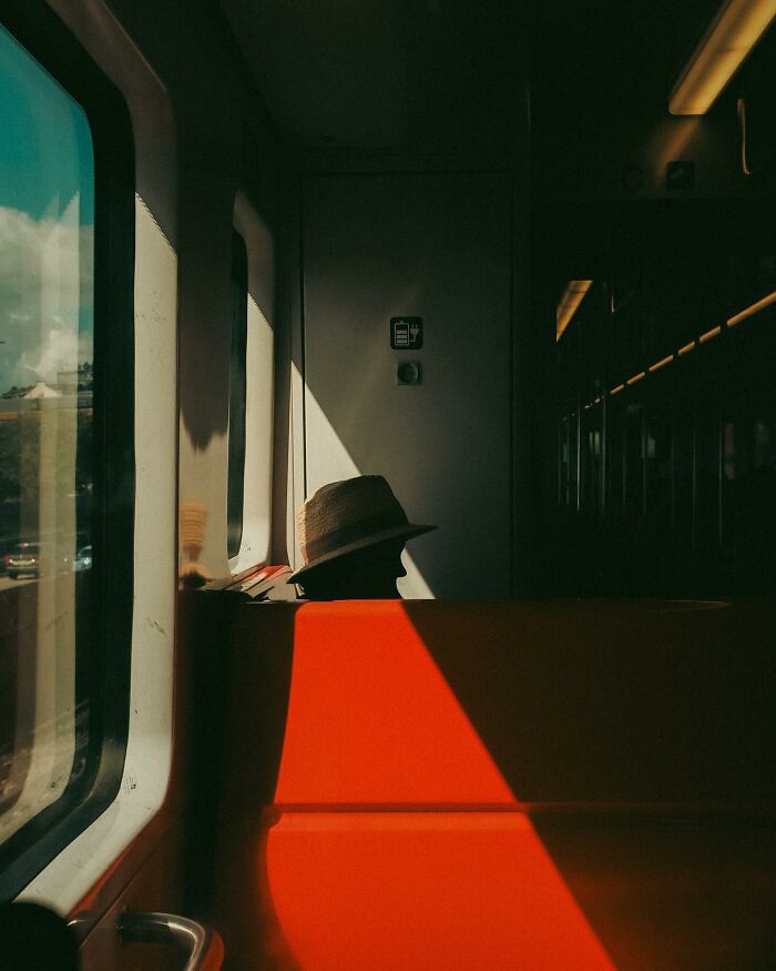Urban photo of a person wearing a hat sitting on a train seat with dramatic lighting and shadows, street moment captured.