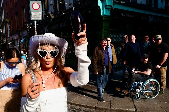 Woman wearing heart-shaped glasses and a white hat, holding an eggplant on a busy street, captured by street photographer.
