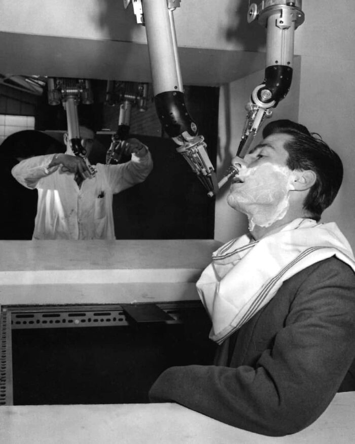 Black and white photo of a man shaving with a robotic arm, a confusing historical invention from the past.