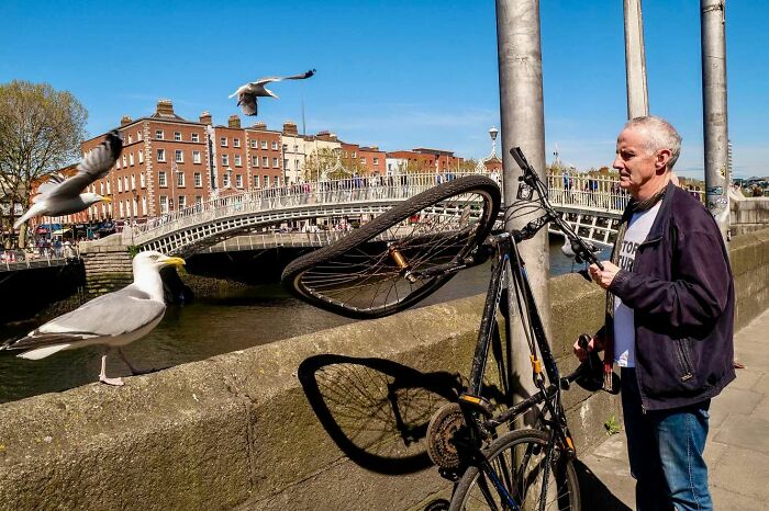 Street photographer capturing a man locking his bike near a river with seagulls flying and a bridge in the background.