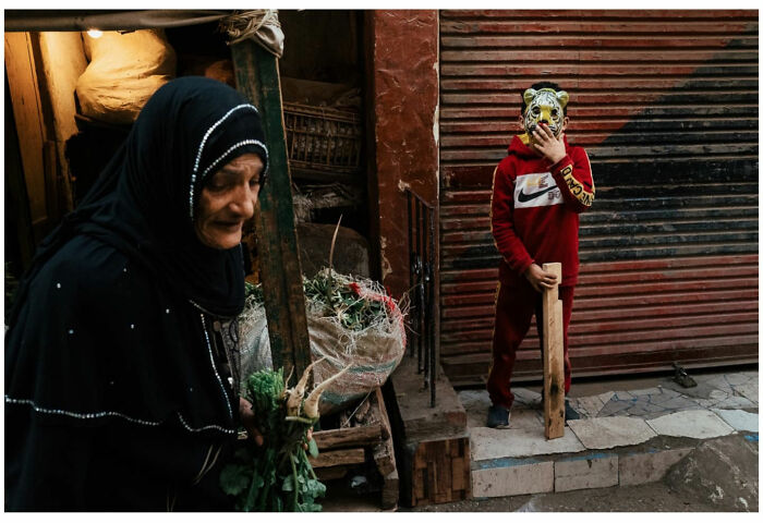 Elderly woman in black hijab holding herbs beside a child in a tiger mask on a street, capturing raw street moments Asia.