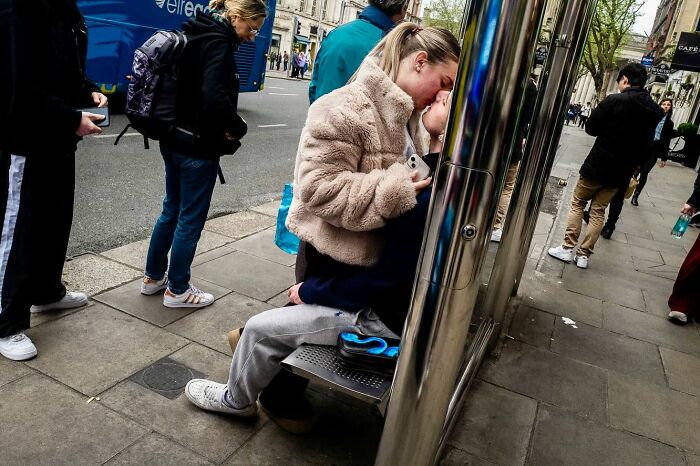 Couple sharing a kiss on a city street bench, captured candidly by a street photographer amidst busy pedestrians.