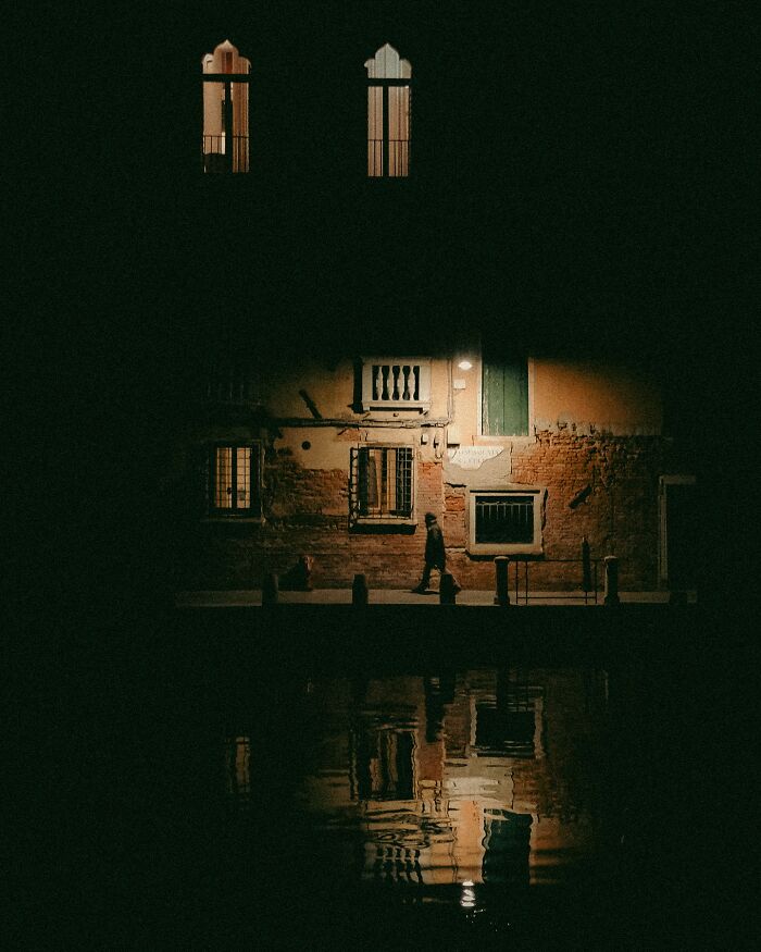 Urban photo of a person walking by a dimly lit building reflected in water, a real moment caught on the street at night.