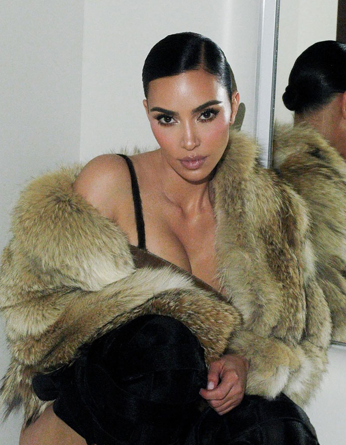 Kim Kardashian wearing a fur coat posing in front of a mirror with discussions about plastic surgeries around her.
