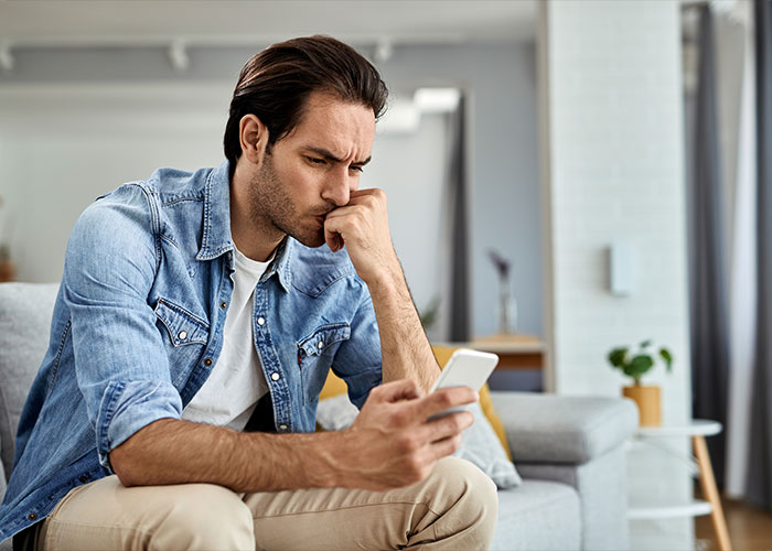 Man in denim shirt sitting on couch, looking thoughtfully at his phone, reacting to creepy facts that stick in the brain.