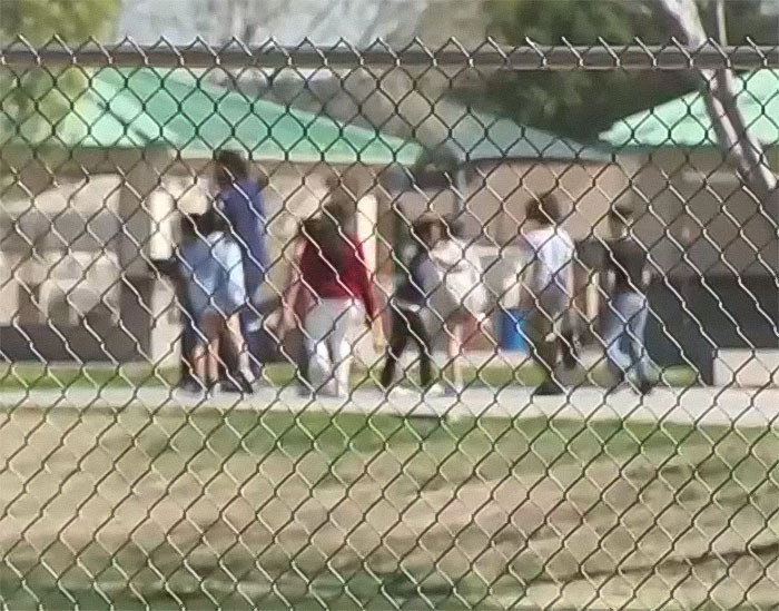 Children playing inside a fenced park area with picnic shelters visible in the background on a sunny day.
