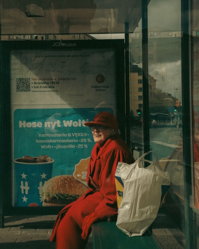 Elderly woman in red coat and hat sitting at a bus stop in Helsinki, urban photos capturing real street moments.
