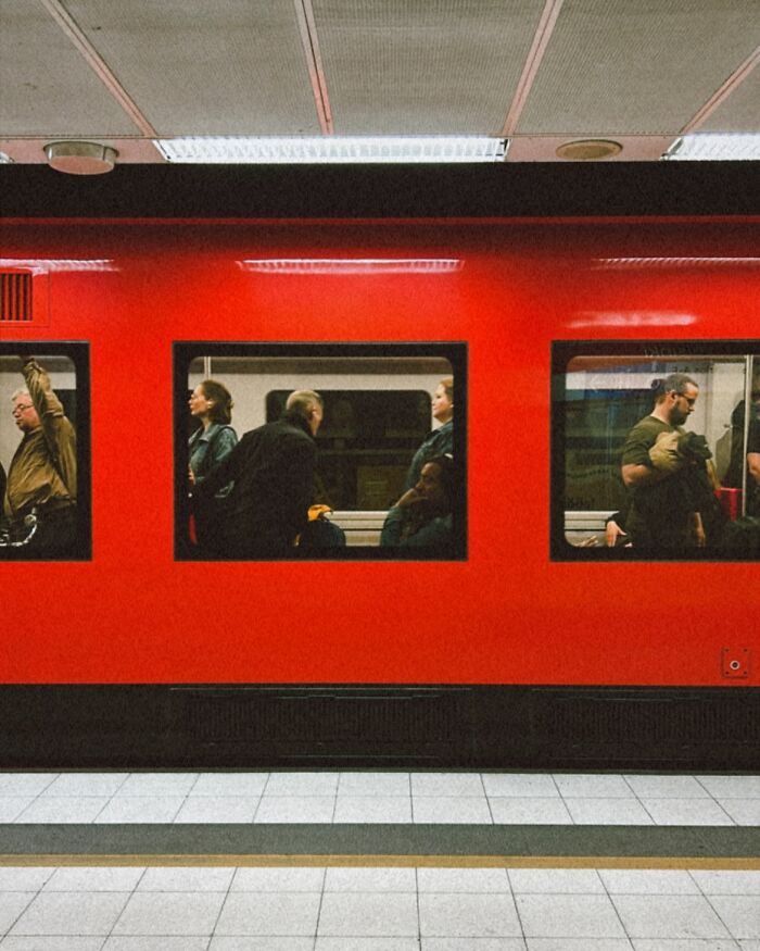 Passengers visible through windows of a red train at a station captured in an urban street photography moment.