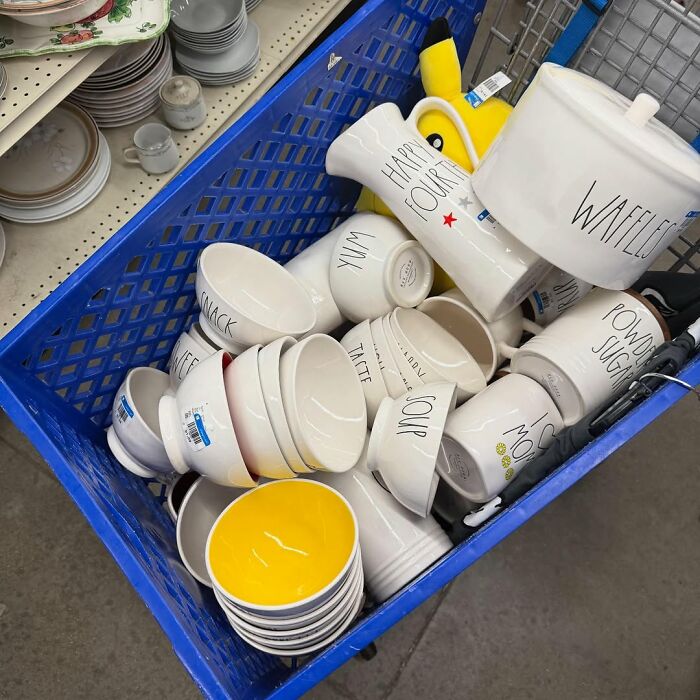 Blue shopping cart filled with various secondhand ceramic bowls, mugs, and kitchenware with assorted labels and designs.