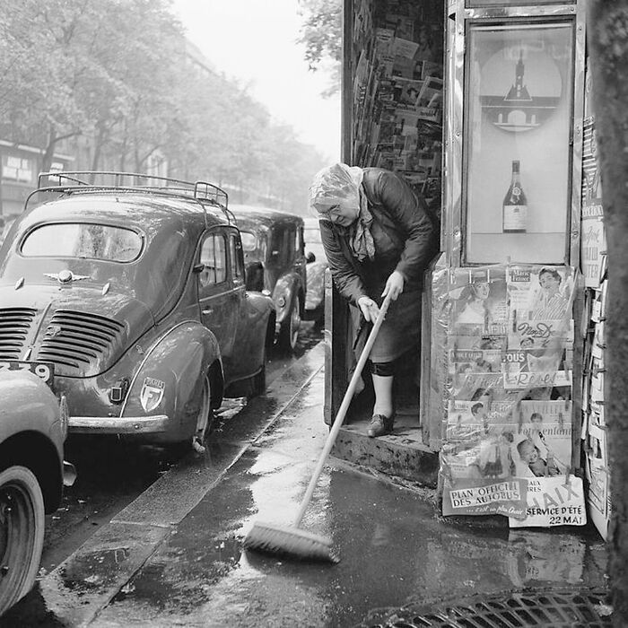 Woman sweeping outside a shop on a rainy street, vintage cars parked, colorized historical photo by viral artist.