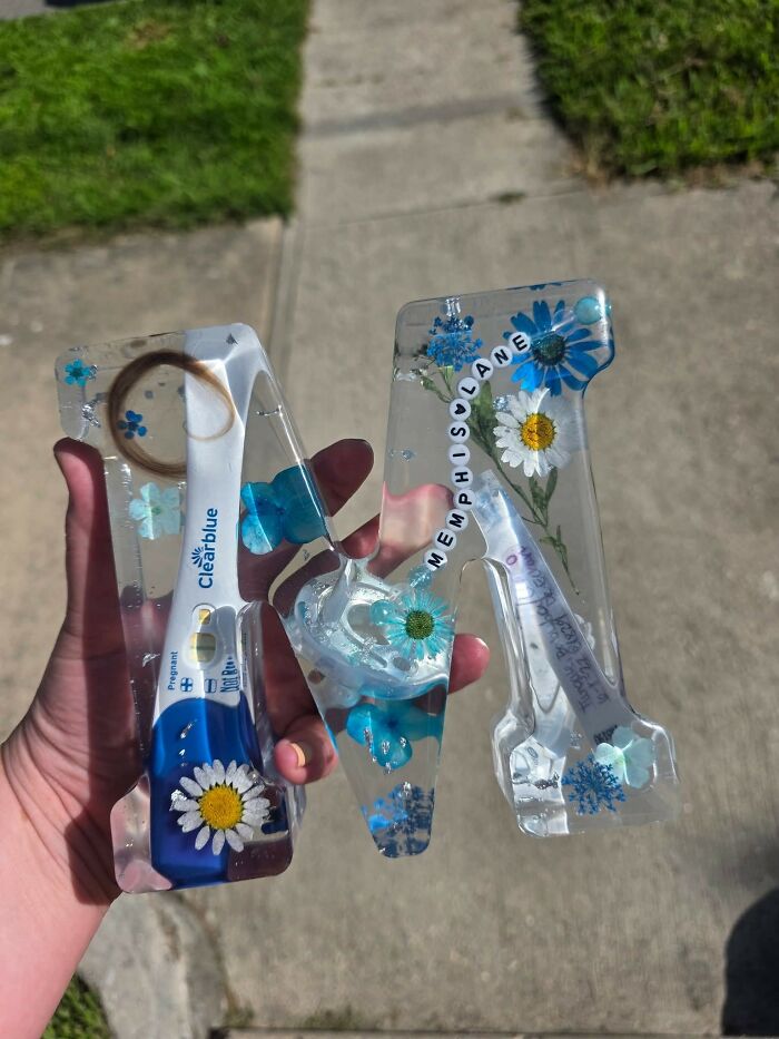 Hand holding resin craft letters with embedded flowers, pregnancy test, and beads, showcasing unusual and questionable crafts.