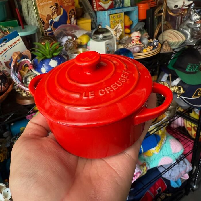 Small red Le Creuset pot held in hand among various cool secondhand finds on a cluttered vintage shelf.