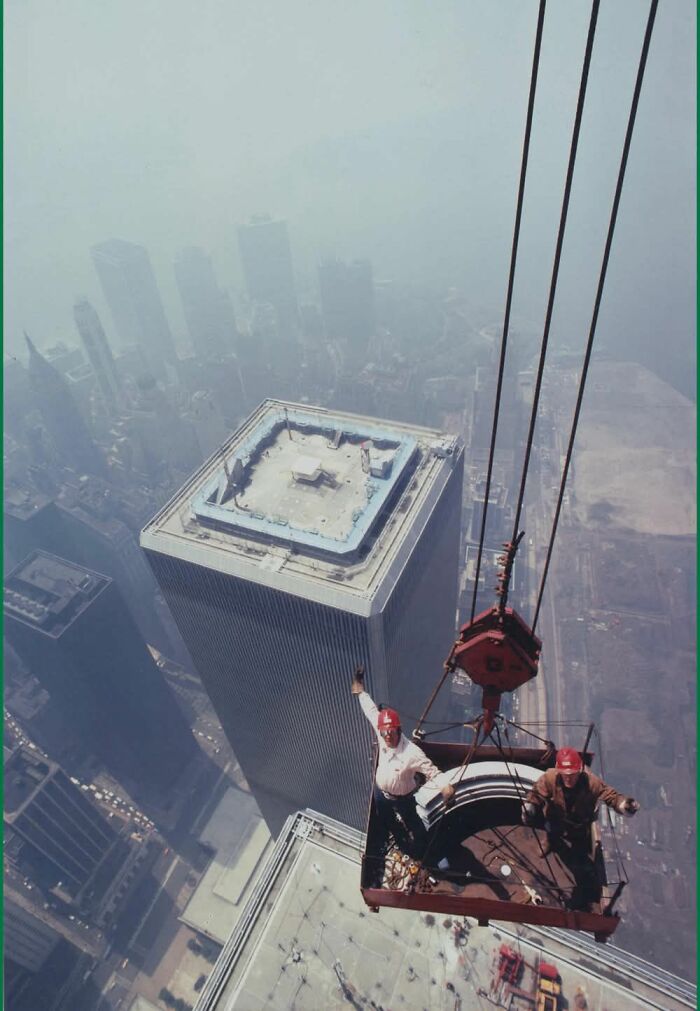 Two construction workers wearing red helmets on a hanging platform high above city skyscrapers historic photo