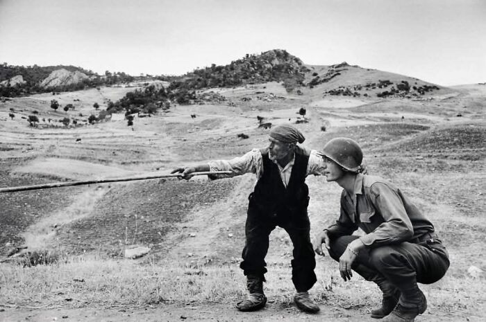 Black and white historical photo of a soldier and an old man in a rugged landscape, capturing a moment from the past.