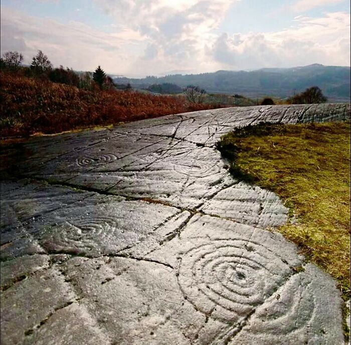 Ancient carved stone spirals and lines etched into a rock surface in an open archaeological world landscape.