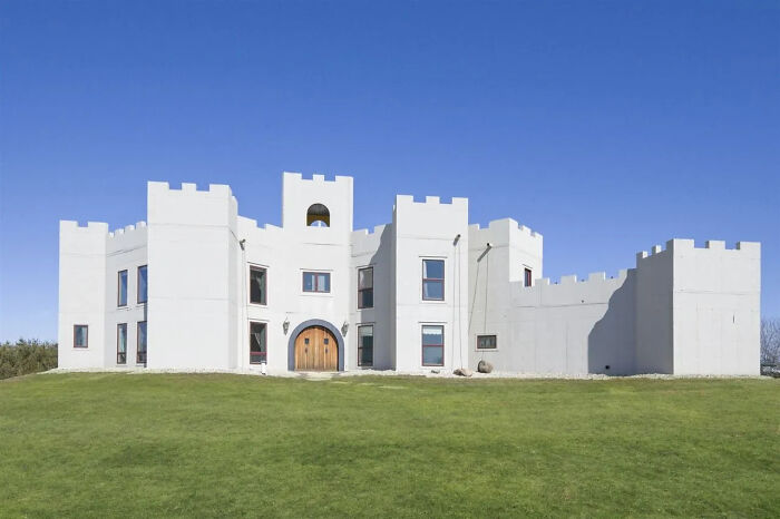 Modern white castle-style mansion with battlements and wooden door on a large green lawn in real estate listings.