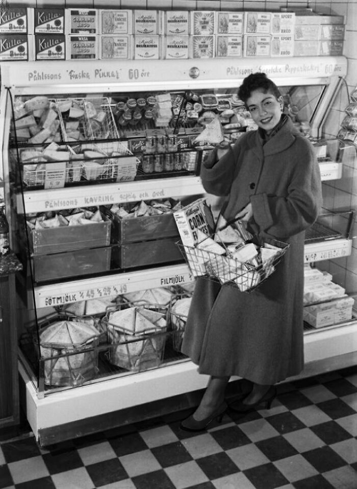 1940s housewife smiling while grocery shopping, holding a basket filled with food items in a vintage store setting