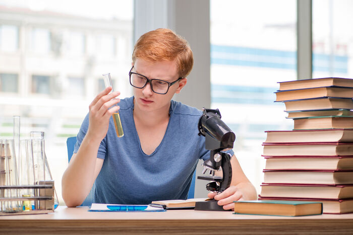 Young man committed to a lie, examining a test tube in a lab setting with microscope and stacked books nearby.