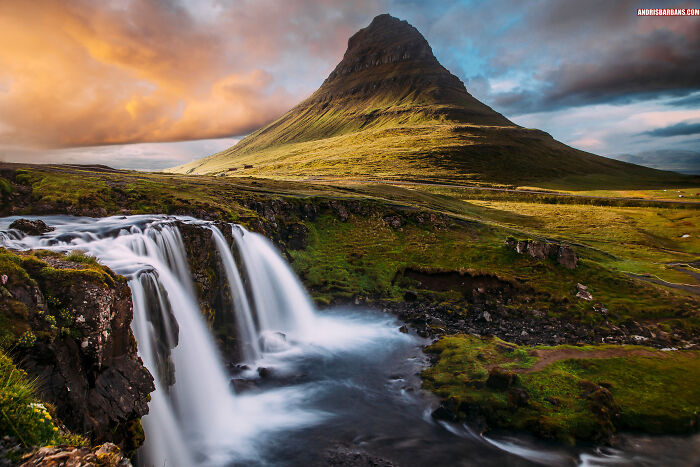 Scenic travel photo of a waterfall and green mountain landscape under dramatic clouds, breathtaking travel photography.