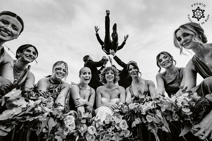 Bride and bridesmaids smiling with bouquets as groomsmen playfully toss a person in the air at a wedding celebration.