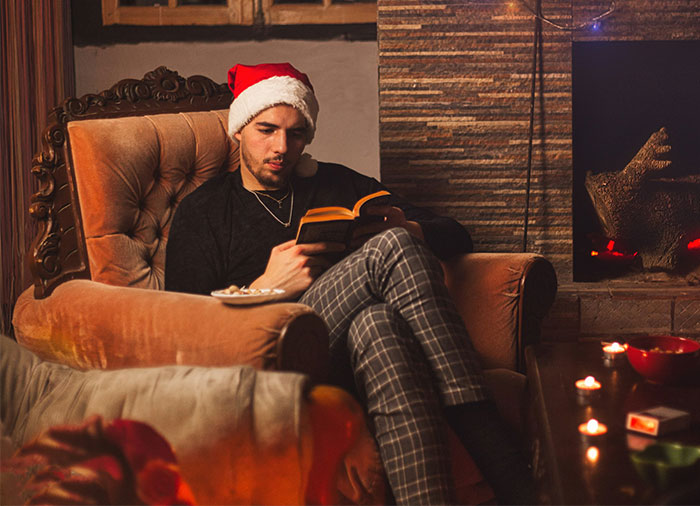 Man wearing Santa hat reading a book by the fireplace, relaxing and using all PTO before project deadline.