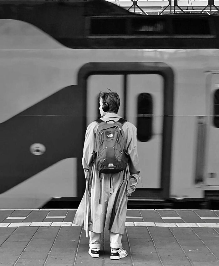 Person wearing headphones and a long coat stands on a platform waiting as train passes in a beautiful street shot.