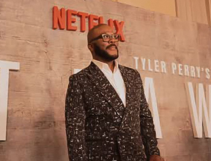 Tyler Perry standing at a Netflix event, wearing a patterned suit and glasses, with promotional background behind him.
